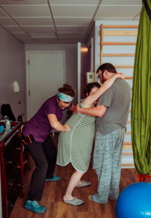 pregnant woman wearing green gown, leaning on husband who is standing. Midwife behind woman, doing hip squeezes through contraction